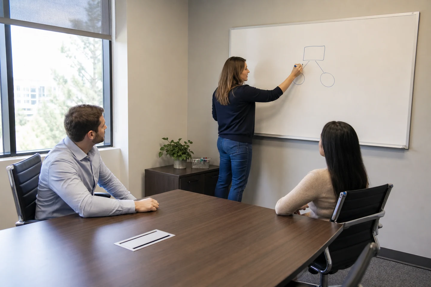 A woman writes a diagram on a whiteboard while two coworkers sit at a conference table.